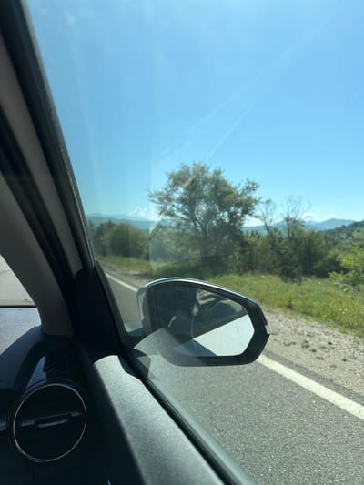 The image shows a view from inside a vehicle, likely a car, traveling on a road in what appears to be a rural or semi-rural environment. The road has a white line marking. A side view mirror of the car is visible. Outside the car window, the scene includes vegetation (trees and shrubs), and hills or mountains in the distance. The sky is blue with no clouds visible. An air vent and part of the car's dashboard are visible in the foreground. The city of Negushevo, Bulgaria is not directly visible within the frame, but it is asserted as the location in the prompt.