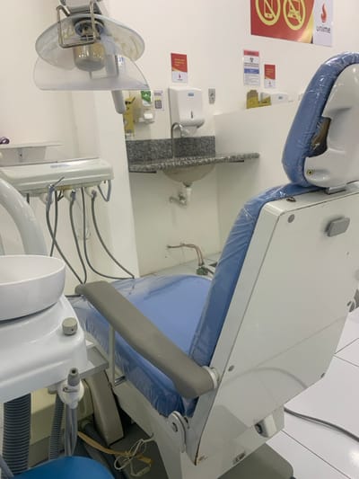 The image displays an unoccupied dental operatory located within a facility associated with Unime, specifically in Lauro de Freitas, Brazil. The room features white tiled flooring and plain white walls.
The central object is a white dental chair, positioned with its backrest and seat upholstered in light blue, both protected by a clear plastic covering. The chair has gray armrests. To its left, a white dental unit includes a circular ceramic cuspidor bowl and an array of grey hoses connected to various dental tools and suction lines. A dental light, equipped with a clear, curved protective shield, is suspended above the chair.
Against the rear wall, a small, light-colored circular sink with a metal faucet is integrated into a grey speckled granite countertop. Above the sink, a white wall-mounted soap dispenser is visible. Multiple signs are affixed to the wall, including a prominent red and white sign featuring the "unime" logo alongside red icons indicating prohibitions such as cell phones and laptops. Another smaller sign displays a biohazard symbol.
No individuals are present in the scene. The equipment appears clean and prepared for use, consistent with a clinical or educational dental environment.