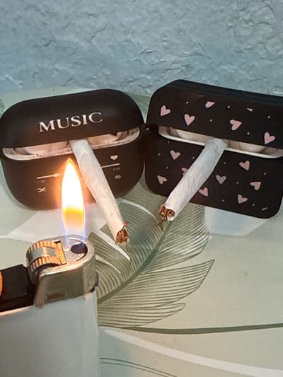 The image depicts a close-up, indoor scene. In the foreground, a metal lighter is actively igniting a flame. To the right of the lighter, two hand-rolled cigarettes are positioned. One cigarette is being lit by the flame from the lighter, while the second cigarette rests adjacent to it. Behind the cigarettes are two black cases for wireless earbuds. The case on the left is open, revealing a glimpse of the earbuds inside, and has the word "MUSIC" printed on its lid. The case on the right is closed and decorated with small pink hearts and dots. The subjects are placed on a surface with a green, feather-like pattern. The background consists of a textured, light blue wall. There is no indication of the city of Nauen, Germany, or any specific location within it.