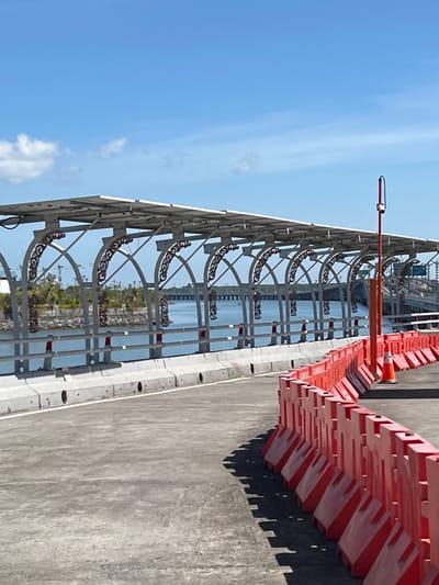 The image shows a bright, sunny day with a clear blue sky and scattered clouds. In the foreground, a grey asphalt road curves to the right. A bright orange plastic barrier with red and white reflective stripes runs along the right side of the road, creating a temporary traffic management system. A traffic cone is visible behind the barrier.
In the mid-ground, a pedestrian walkway or bridge is visible. It is covered by a structure with solar panels on top, supported by metal beams and decorative arches. The walkway has a grey concrete barrier along its edge, with circular openings visible at intervals. Beyond the walkway, a body of water is visible, with a bridge extending across it in the distance. Lush green vegetation and trees can be seen on the shore to the left of the walkway.
The scene is likely the **Benoa Bridge** in South Denpasar, Bali, Indonesia, which is known for its distinctive solar panel-covered walkway and its location over a bay. The presence of construction barriers suggests ongoing work or maintenance. No people are visible in the image. The overall impression is one of a modern infrastructure project in a coastal environment.
