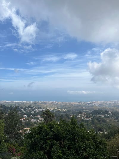 The image depicts a daytime view from a high vantage point, possibly in Mijas, Spain, looking out over a cityscape descending down a hillside towards the coast. The sky is partly cloudy, with blue sky visible among the white and grey clouds. The terrain below is densely covered in greenery, interspersed with white buildings and houses that suggest a residential area on the outskirts of the city. In the foreground, dense vegetation and trees partially obscure the view, indicating the perspective is from within a garden or wooded area. The coastline is visible in the distance, merging with a hazy atmosphere. There are no people visible in the frame.
