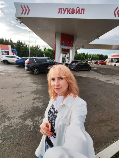 A woman with blonde, wavy hair is taking a selfie at a Lukoil gas station in Staraya Yum'ya, Russia. She is wearing a white open shirt over a black and white patterned t-shirt, blue jeans, and a ring on her left hand. Her fingernails are painted red. Behind her, several cars are parked at the gas pumps under a white canopy with the Lukoil logo visible. In the background, there are trees, a cloudy sky, and other buildings associated with the gas station, including a small convenience store. One of the gas pumps shows prices for different fuel types.