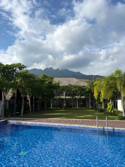 This is a landscape photograph of a swimming pool and resort area. In the foreground, a bright blue swimming pool with tiled flooring dominates the lower half of the image. The water is disturbed, creating reflections and patterns. A wooden deck surrounds the pool, with two silver handrails for pool access. Beyond the pool and deck, a well-maintained lawn stretches towards a row of buildings. These buildings appear to be part of a resort, with thatched roofs and multiple levels. Lush green trees, including palms, are scattered throughout the property, providing shade and a tropical atmosphere. In the background, a range of mountains looms under a sky filled with dramatic, white clouds. The overall scene suggests a serene and relaxing resort environment in Pasir Putih, Indonesia. There are no people visible in the image.