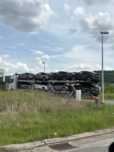 The image shows a car carrier truck filled with black Citroen C4 cars parked next to a roadside in Aubange, Belgium. The truck is oriented perpendicular to the road. The scene includes a cloudy sky, a green hill in the background, a light post, a green grassy verge, and a section of a concrete road barrier with a drainage grate. A fuel station sign is visible in the lower left corner. No people are apparent in the image.