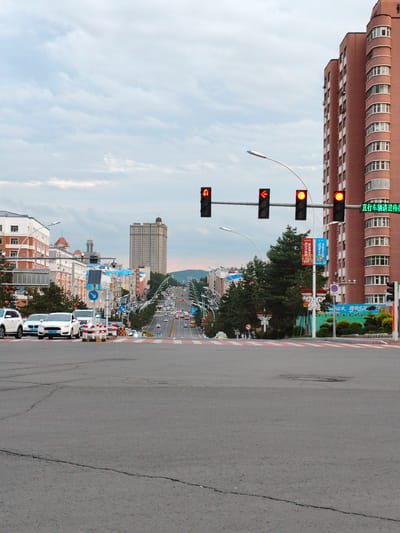 The image shows a street view in Jixi, China, likely during the daytime with overcast skies. The foreground features a wide asphalt road with a marked pedestrian crossing in red and white stripes. A few cars are stopped at the crossing. In the background, a long, uphill road stretches into the distance, lined with buildings on either side and trees. Traffic lights are visible, with some showing red. The architecture includes modern apartment buildings and older structures. The overall scene depicts a typical urban street in a Chinese city, with elements of urban infrastructure and daily life.