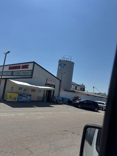 A low-angle, exterior, daytime shot captures a commercial building with a large silo structure in the background, under a clear blue sky. The building, identified by red lettering as "КАРАОВО МЕС," appears to be a retail establishment. In front of the building, several freezers display "ICE CREAM" advertisements. To the right, two cars, a dark hatchback and a silver wagon, are parked. The area is paved with interlocking bricks in the foreground, transitioning to asphalt. The scene is likely located in or near Vedrare, Bulgaria, given the presence of Cyrillic script.