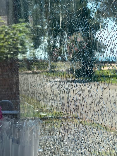 The image shows a close-up view of cracked glass, possibly a window or windshield. The glass is fractured into a dense network of fine lines. Through the cracked glass, a blurred outdoor scene is visible. This scene includes green foliage, trees, a pathway or road, and a body of water in the distance, possibly the sea. The lighting suggests it is daytime. In the foreground, to the left, a blurred object with white elements and some pink color is partially visible, which might be part of a display or arrangement. The overall impression is that the photograph was taken from inside looking out through damaged glass.