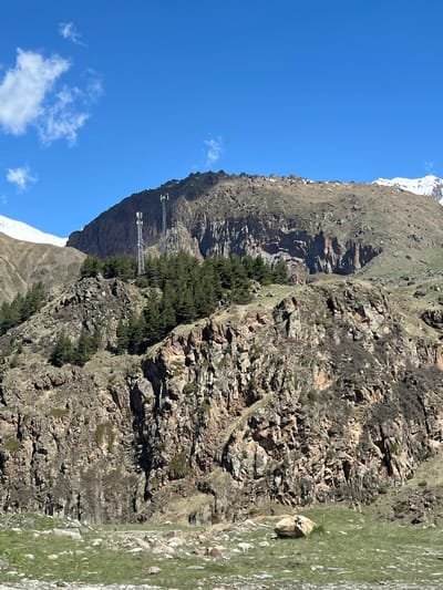 The image depicts a mountainous landscape under a clear blue sky with scattered clouds. The mountain's terrain is rugged, with exposed rock formations and sparse vegetation. Patches of green grass and some trees are visible on the slopes. In the distance, there is what looks like a structure with cables for ski lifts, on the side of the mountain. The location may be in Sioni, Georgia, based on the user's information.