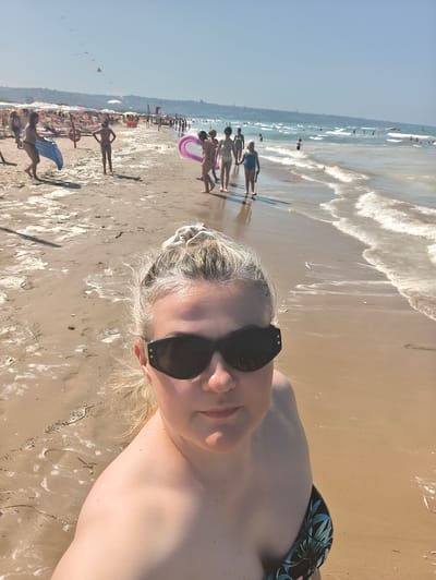 The image is a selfie taken from a low angle on a sunny day at the beach in San Salvo, Italy. The foreground features a woman wearing sunglasses and a strappy bikini top with a tropical leaf pattern. Her blonde hair is pulled up in a messy bun. The background shows a crowded beach with numerous people in swimwear, many walking along the wet sand near the water's edge. Beach umbrellas, loungers, and inflatable toys are visible. The sea is calm with gentle waves and clear blue skies. The sand is light brown and appears wet and somewhat foamy in patches, suggesting the tide has recently receded. Some kites are flying in the distance.