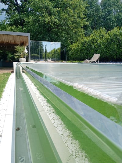 The image depicts an outdoor swimming pool area in Nowa Wieś Szlachecka, Poland. The pool is covered with a transparent, slatted material, and the water within is a vibrant green color. A silver-colored handrail runs along the edge of the pool. To the left of the pool, there is a white concrete border filled with white pebbles and some greenery. Further left, a grassy area is visible. In the background, a modern structure with a thatched roof and a potted palm is partially visible, along with a large mirrored wall reflecting the surroundings. Behind the mirrored wall, a light-colored lounge chair sits on the pool cover. A dense row of green bushes borders the back of the scene, with a thick canopy of mature trees providing shade. The sky is partly cloudy. There are no people visible in the image.