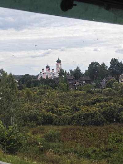 The image depicts a pastoral scene with a prominent pink and white church topped with dark domes, situated in the distance. The church is surrounded by dense green foliage and a cluster of rustic wooden houses. The foreground features a field of tall green grass and wildflowers. The sky is overcast with clouds, suggesting a temperate climate. The scene appears to be a settlement within or near Ямская Слобода, Russia, characterized by its traditional architecture and natural landscape.