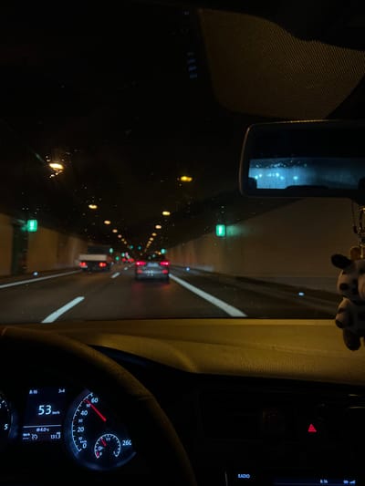 The image captures a first-person perspective from inside a car driving through a tunnel at night. The view is primarily through the front windshield, which appears to have raindrops or condensation on its surface.
In the foreground, car interior elements are visible. On the right, a portion of the steering wheel and the instrument cluster are seen. The speedometer indicates a speed of "53 km/h," and the digital display shows "8:16" (likely the time), an outside temperature of "4.0°C," and a mileage reading of "1313." Above the instrument cluster, part of a radio unit is visible with the word "RADIO" and the time "8:16" again. The top center features a rearview mirror, from which a small, black-and-white spotted plush toy (resembling a cow or dalmatian) hangs.
The main scene outside the car is a dark tunnel, illuminated by rows of lights. Yellow/orange lights line the left wall, while green and red lights, presumably from other vehicles, are visible further down the tunnel. The tunnel's walls appear to be a neutral grey, with some reflective vertical lines. The ambient darkness and reliance on artificial lighting suggest either nighttime driving or very dim conditions within the tunnel. The "km/h" for speed and "°C" for temperature are consistent with the provided location context of Germany.