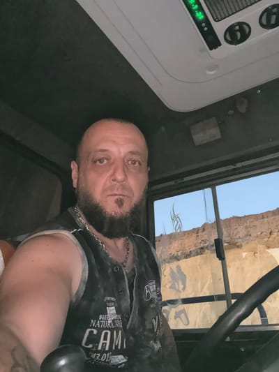 The media file shows a man in the driver's seat of a vehicle, looking at the camera. He has a shaved head, a beard, and is wearing a dark vest with white trim over a grey undershirt. A silver chain is visible around his neck. The man's expression is neutral.
The interior of the vehicle is dark, with a white console on the left side displaying a digital readout. Above the man's head, there is a sunroof or window that looks out onto a desert-like landscape with rocky cliffs under a clear blue sky. The sun appears to be low in the sky, suggesting either sunrise or sunset.
On the window, there are dark tribal-style decals. The man's vest has text printed on it, including the word "CAMP" prominently displayed, along with other words that are difficult to read but appear to be in English, such as "NATURAL ARE" and "DEPARTMENT." The man's arm is resting on something, possibly the armrest of the seat.
The scene is likely taking place in a rural or remote area, given the natural landscape visible through the window. The lighting indicates it is either early morning or late evening.