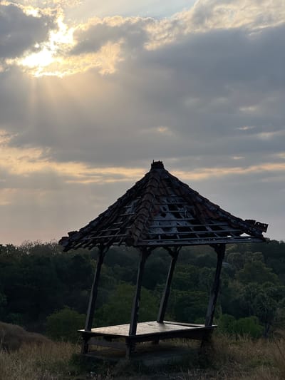 An abandoned open-air structure sits on a grassy knoll, silhouetted against a cloudy sky at sunset in Kubutambahan, Indonesia. The structure, possibly a gazebo, has a weathered, tiled roof with missing sections, revealing the wooden beams beneath. The wooden platform appears damp, reflecting the fading light. Lush green trees form a dense backdrop to the structure, with tall grasses in the foreground. Sunbeams break through the clouds, casting a warm glow on the scene. The image captures a serene yet slightly melancholic atmosphere.