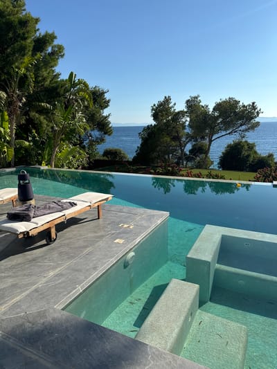 The image depicts a luxurious infinity pool overlooking the Aegean Sea, likely situated in the Vouliagmeni Municipal Unit of Greece. The pool is a prominent feature, with its turquoise water reflecting the clear blue sky and lush green trees. In the foreground, a modern lounge chair with a white cushion and folded grey towels sits on a grey stone deck. A black portable speaker is placed on the lounge chair. The pool's edge is lined with stone, and steps descend into the shallower end of the pool, where small fish are visible. Beyond the pool, a well-maintained lawn is bordered by vibrant red flowering bushes and tall pine trees. The sea stretches out to the horizon, with distant landforms visible under a cloudless sky. The scene exudes tranquility and opulence, suggesting a high-end resort or private residence.