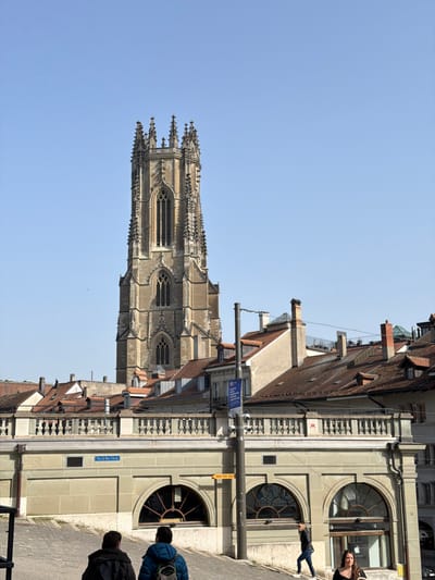 The image displays an urban street scene in Fribourg - Freiburg, Switzerland, focused on the St. Nicholas Cathedral (Cathédrale St-Nicolas) tower. The sky is clear and blue.

The dominant feature is the tall, ornate Gothic bell tower of St. Nicholas Cathedral, constructed from light-colored stone, positioned centrally in the background. It exhibits multiple levels of arched windows, intricate tracery, spires, and pinnacles.

Below and to the right of the cathedral, a dense collection of buildings with traditional reddish-brown tiled roofs and numerous chimneys extends across the mid-ground. In front of these structures, a multi-story, light-colored building is present, featuring a balustraded terrace at its top. Its lower level includes three large arched windows, one of which displays an Apple logo inside. A sign reading "Place de Notre-Dame" is affixed to the building's facade, and a yellow sign with "Hôtel Ville Ardou" is also visible. A dark utility pole stands vertically in the mid-ground, partially obscuring the building on the right.

The foreground depicts a paved, upward-sloping street or pathway. Three individuals are visible on this path. Two individuals, seen from behind, walk uphill: one on the left in a dark jacket, and another in the center wearing a blue jacket and a backpack. A third individual, a female with dark hair, walks downhill on the right, wearing a dark top and light trousers, carrying a bag. A fourth individual, a male in a dark jacket and jeans, is visible further up the slope on the right, also walking downhill.