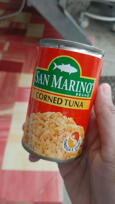 A hand holds a can of "San Marino Corned Tuna" in an indoor setting, possibly a home or store. The can is red with green and yellow accents, featuring a white fish logo and text. The tuna inside is visible at the bottom of the can, appearing flaky and light brown. A label with "Omega 3 DHA" is also present. The background is out of focus, showing a patterned surface that could be a tablecloth or floor. The image does not provide any specific information to determine its location within Kawit, Philippines.