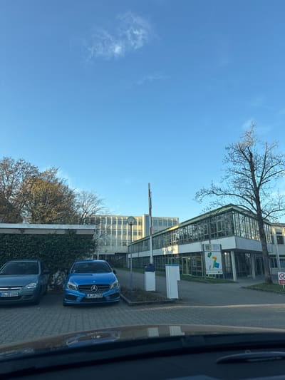 The image depicts an outdoor parking area and buildings at the Robert-Schuman-Schule in Bühl, Germany, specifically at Steinfeldstraße 10, 77815 Bühl. The scene is observed from within a vehicle, with its dashboard or hood partially visible at the bottom of the frame.
Under a clear blue sky with a few wispy white clouds, the primary subjects are institutional buildings and parked vehicles. On the left, a light blue/grey Opel Corsa D (license plate "RA JD 2008") and a blue Mercedes-Benz A-Class (license plate "LR VM 1708") are parked. Behind these vehicles, a low structure covered with dense green ivy is visible, backed by deciduous trees with varying foliage, some bare.
In the center, a white and red striped barrier arm is upright, indicating an open entry or exit. Two white vertical bollards or barrier mechanism housings stand near it. To the right of the barrier, a multi-story building with a distinctive sloped glass roof section and large windows forms a prominent part of the midground. A tall, bare deciduous tree stands to the far right of this building. A large informational sign with colorful geometric elements (green, yellow, blue) and white text, including "HAUPTEINGANG" (main entrance), is positioned in front of the glass-fronted building. A red and white "no parking/stopping" sign is partially visible on the extreme right.
In the background, behind the glass-fronted structure and barrier, a larger, multi-story building with numerous windows and light-colored facade panels is visible. A large circular clock or light fixture is affixed high on its exterior. No individuals or active interactions are visible in the static scene.