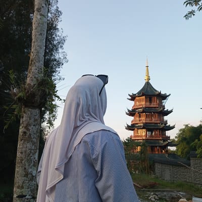 The image captures a scenic view of a tiered, traditional East Asian pagoda with a golden spire, illuminated by warm sunlight, located in the city of Suka Makmur, Indonesia. In the foreground, a person wearing a light purple hijab and a blue and white striped shirt faces away from the camera, looking towards the pagoda. The person has dark sunglasses perched on top of their head. To the left of the frame, a thick, gray tree trunk, partially covered in lichen and a small cluster of plants, stands prominently. The background features lush green foliage and a clear blue sky. A stone wall and more vegetation are visible near the base of the pagoda, suggesting it is situated within a well-maintained garden or cultural site. The lighting indicates it is likely late afternoon or early morning, given the warm glow on the pagoda.