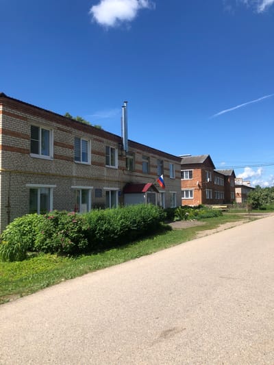 The image depicts a row of low-rise brick buildings in Izdeshkovo, Russia. The buildings are of varying colors, with some being a light beige and others a darker red brick. They appear to be multi-story residential or administrative structures, featuring numerous windows. A Russian flag is visible on one of the buildings. Green bushes and trees line the front of the buildings, and a paved road runs in the foreground, leading away from the viewer. The sky is blue with scattered clouds.