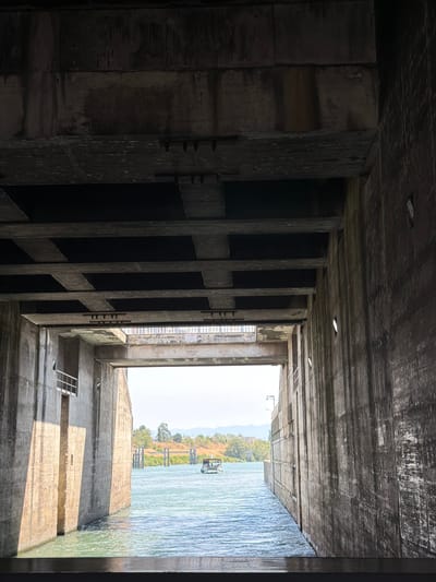 The image shows a view from within a concrete structure, looking out towards a body of water. The structure has rough, textured concrete walls and a ceiling made of concrete beams with dark, rectangular panels between them. A railing is visible on the left side of the structure.
The water is a bright blue-green color and appears to be flowing. On the water, in the distance, is a small white boat with several people on it. The boat is moving away from the viewer.
Beyond the water, there is a shoreline with trees and some distant hills or mountains under a clear, bright sky. There are some pylons or posts in the water near the shoreline. The overall impression is one of a large, industrial concrete facility opening onto a natural landscape and a waterway.
This scene does not appear to be located in Bourg-lès-Valence, France. The architecture and landscape are not characteristic of that region.