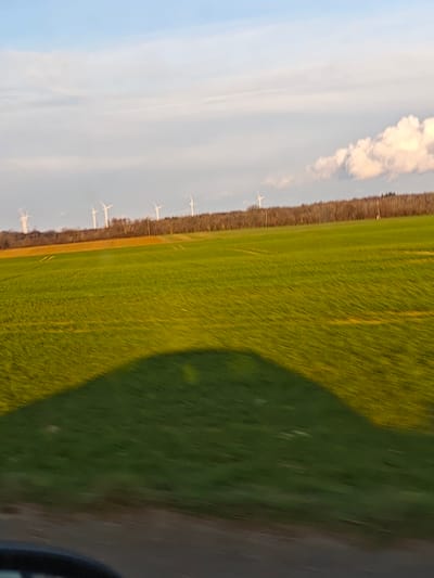 The image displays an expansive agricultural landscape in the vicinity of Aumetz, France, captured from a moving vehicle, indicated by motion blur across the foreground and midground. In the immediate foreground, a dark, partially visible object resembling a vehicle's side mirror is present at the bottom left edge, with a large, dark shadow, consistent with a vehicle, extending diagonally across the lower-left portion of a vibrant green field. This field, likely planted with young crops, occupies the majority of the midground, showing faint linear patterns from cultivation. To the left, behind the green field, a section of a contrasting yellowish-brown field is visible. A dense, horizontal band of leafless deciduous trees forms the background, separating the fields from the sky. Above this tree line, approximately eight white wind turbines with visible blades are dispersed, suggesting a wind farm. The sky is a light blue with scattered white and grey clouds, including a more prominent, brighter white cloud formation on the right. The lighting, characterized by a golden hue on the field and the long shadow, suggests late afternoon or early morning. No individuals or specific interactions are visible within the frame.