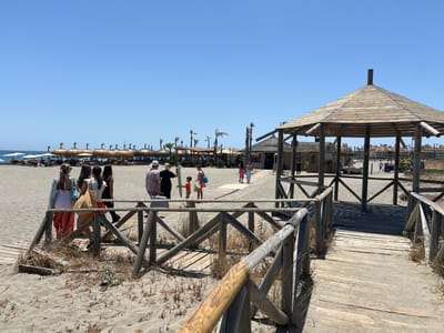 This is a photograph of a beach in San Roque, Spain, on a clear, sunny day. The foreground features a wooden boardwalk with railings, leading onto a sandy beach. Several people are visible on the boardwalk and the beach.
To the left, a group of four women are walking on the boardwalk, some carrying bags. One woman wears a white top and a long, colorful skirt. Another wears a white top and orange shorts. Further back, a man is walking away from the camera on the beach, and a young boy in an orange shirt is walking towards him. Another woman in a pink dress and a blue bag is walking behind the boy.
In the mid-ground, a rustic, hexagonal wooden gazebo with a thatched roof stands to the right, providing shade. Behind it, a beach bar or restaurant with a thatched roof is visible. Further into the background, rows of straw-covered beach umbrellas with lounge chairs are set up on the sand. Palm trees are scattered throughout the scene. Beyond the beach, buildings with terracotta roofs are visible in the distance, suggesting a coastal town or resort. The sea is not directly visible in this frame, but the presence of the beach umbrellas and lounge chairs indicates it is nearby. The sky is a clear, bright blue.