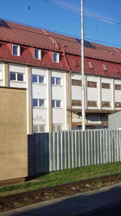 A wide shot captures the side of a long, white, multi-story industrial building with a red-tiled, sloping roof. The building features several rows of windows with white frames. The roof has dormer windows and some solar panels visible on the right side. A tall, silver utility pole with crossbars stands in front of the building, obscuring a portion of its facade. In the foreground, a corrugated metal fence runs horizontally across the image, separating the building from a patch of green grass and railway tracks. The sky above is a clear, bright blue with a single bird in flight. The overall scene depicts a typical urban industrial area. The image was taken in the city of Spišská Nová Ves, Slovakia.