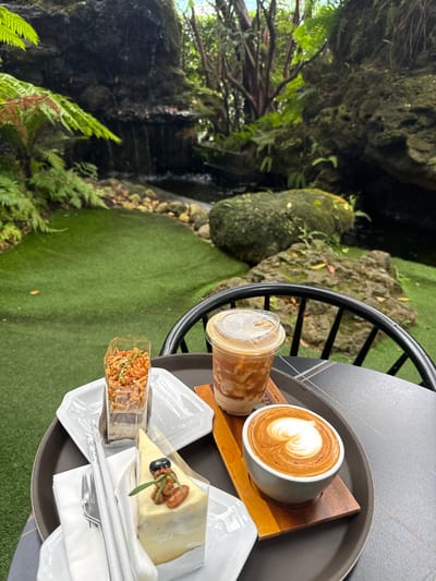 A tray of coffee and desserts sits on a dark, round table in an outdoor seating area. The tray holds a cup of latte art coffee, a clear plastic cup with an iced coffee drink, a slice of white cake topped with a blueberry and nuts, and a slice of chocolate cake with almonds. The table is positioned in front of a lush, green artificial lawn area, which leads to a rocky landscape with a small waterfall and dense foliage. The scene appears to be set in a cafe or restaurant located in Ban Na Chom Thian, Thailand, emphasizing a serene and naturalistic ambiance.
