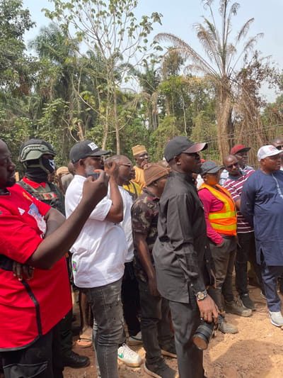 A group of approximately ten men are gathered outdoors under bright daylight in what appears to be a rural or semi-rural area in Aloko, Nigeria. The image captures them from a low angle, standing on packed dirt or sand, with dense green foliage and palm trees forming the background. The sky is clear, indicating sunny weather.
The men are dressed in various casual attire. Notable clothing includes a man wearing a bright orange safety vest over a pink/red shirt, another in a white shirt, one in a black shirt and black cap, and another in a blue long-sleeved top. Several men are wearing caps; one black cap has "SN" or "SIT" visible, and a white cap in the background shows partial text like "C_MP_SS".
Many of the men are looking upwards or forward, suggesting they are observing an event or object outside the camera's frame. One man in a black shirt is holding a professional camera with a large lens, pointing it forward. Another man in a white shirt is holding a smartphone, seemingly recording or taking a picture. The collective gathering, coupled with the presence of cameras and a safety vest, suggests they might be attending an inspection, a community event, or documenting a happening in the area. No strong emotions are clearly discernible; most appear focused on what they are observing.