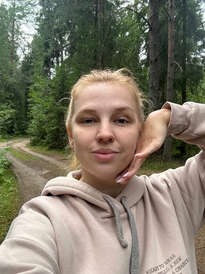 The image shows a young woman with blonde hair and freckles taking a selfie outdoors. She is wearing a light-colored hooded sweatshirt. Her right hand is resting on her cheek, and she is looking at the camera with a slight smile. The background features a dirt road winding through a dense forest with tall trees. The location is specified as Anteplevo (Okhotinskoe S/P), Russia.