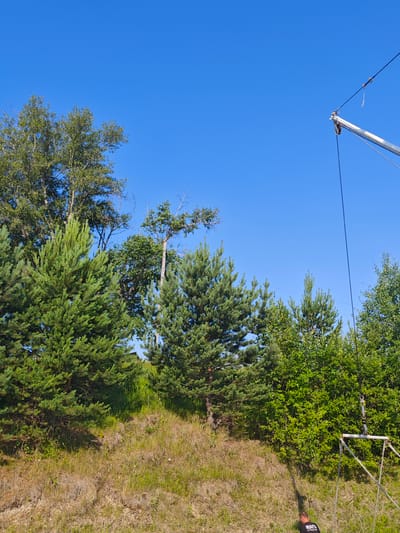 The image shows a clear blue sky above a grassy slope with various trees. In the foreground, there are pine trees and other deciduous trees. On the right side of the image, a metal structure with ropes and pulleys is visible, extending upwards into the sky. The structure appears to be part of some kind of equipment or setup. A person wearing a black shirt with white lettering is partially visible at the bottom right of the image. The overall environment suggests an outdoor setting, possibly a rural area or park. The specific location in Galanino, Russia is not visually discernible from the image content itself.
