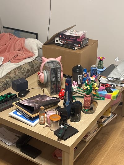 The image shows a cluttered shelf with various objects. There are books, Lego sets, candles, headphones, spray cans, vases with flowers, and other miscellaneous items. A cardboard box is visible on the left side of the shelf. There is no specific information indicating the location in Fulda, Germany.