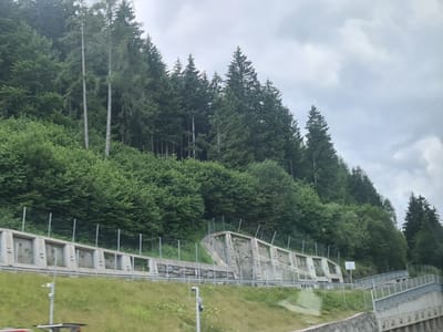 The image captures a view of a steep, forested mountainside in St. Georgen ob Judenburg, Austria, under an overcast sky. The foreground features a constructed embankment designed to prevent rockfalls or landslides. This embankment consists of a series of concrete retaining walls with visible reinforcement. A metal safety fence runs along the top of the embankment.
The slope above the retaining walls is covered with dense green foliage, primarily trees and bushes, indicative of a natural, mountainous environment. The trees are tall and closely packed, with some taller, more slender trees interspersed among the broader deciduous trees.
In the mid-ground, a road or pathway is visible, bordered by a metal railing. There are also several signs and poles present, suggesting infrastructure associated with the road or the mountainside. The overall scene conveys a sense of natural beauty combined with man-made efforts to manage and secure the terrain. No people are visible in the image.