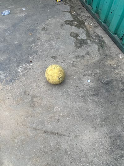 A yellow soccer ball sits on a gray concrete surface. The surface is cracked and dirty, with litter scattered around. A bottle cap rests above the ball. Green corrugated metal is visible at the top left edge of the frame. The lighting is bright, casting soft shadows around the ball and debris.
