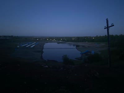 The image depicts a landscape at dusk or dawn, with a body of water, possibly a lake or large pond, as the central element. To the left of the water, there appear to be several white, rectangular structures on the ground. The background features trees and faint lights suggesting a town or city in the distance. To the right of the water, a utility pole stands prominently. The foreground is dark, composed of dirt and rocks, obscuring details. The overall lighting suggests it is either very early morning or late evening. The city of Южный, Russia is mentioned in the prompt as the location.