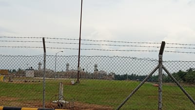 The image depicts an industrial facility, likely a power plant, viewed through a chain-link fence topped with barbed wire. In the foreground, the fence is prominent, with a metal post and diagonal supports visible. Beyond the fence, a grassy field stretches towards the industrial complex. The complex itself features several large structures, including tall smokestacks or chimneys, and numerous pipes and smaller buildings. A lamppost is also visible within the facility grounds. The sky is overcast with clouds. The overall scene suggests a secure, operational industrial area, possibly in Glokaron, Indonesia, given the context. There are no visible people in the image.
