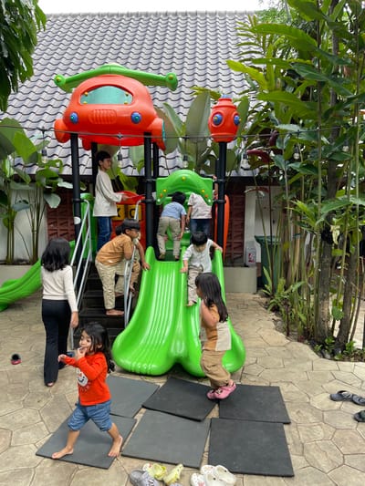 This image depicts an outdoor children's playground located in Garut, Indonesia. The central subject is a multi-component plastic playground structure, predominantly green and orange. It features a bright green double slide, a central staircase with dark grey steps, and an upper platform supporting an orange, stylized helicopter-like section with green rotor blades, and a red cylindrical fixture resembling a fire hydrant.

Eight individuals are present: seven children and one older female or youth. On the playground structure, an older boy in a cream long-sleeve shirt stands on the upper platform, holding a red object. One boy in a brown short-sleeve shirt ascends the dark grey steps barefoot. Two boys, dressed in light blue shirts, climb into an elevated green section of the structure from the right. A boy in a light cream long-sleeve shirt is in the process of descending the central green slide, barefoot. At the base of this slide, a girl with long dark hair, wearing a brown and beige two-tone top and light brown cargo pants, stands in pink Crocs.

On the ground level, a female with long dark hair, wearing a cream long-sleeve top and black pants, stands to the left of the stairs, facing the playground. She is barefoot, with a black and red sandal nearby. In the foreground, a young girl with long dark hair, wearing an orange long-sleeve shirt and blue denim shorts, stands barefoot on a black square rubber mat, her right hand near her mouth.

The ground surface beneath the playground is paved with light brown/grey interlocking stone pavers, interspersed with black square rubber mats. A collection of various colored Crocs and sandals are gathered in the lower right foreground. The playground is surrounded by dense, lush green foliage and trees, particularly on the right side. A building with a dark grey, ridged tiled roof is visible in the background, along with a brown wooden slatted wall. String lights are strung overhead across the top portion of the scene. In the distant background on the right, a partially visible "TOILET" sign, two green trash bins, and a white container are present. The scene is illuminated by natural daylight.
