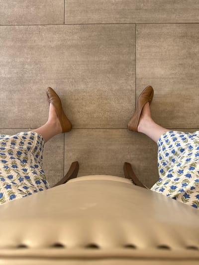 The image shows a person sitting in a chair. The person is wearing a floral print dress and brown leather ballet flats. The chair is white with a tufted back. The floor is tiled. The location appears to be indoors and is unlikely to be identifiable as being in Chula Vista or any other specific location.