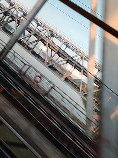 The image shows a view from inside a train carriage crossing a bridge. The train tracks are visible in the foreground, appearing blurred due to motion. Beyond the tracks, a large metal truss bridge spans a body of water. The bridge is made of interconnected steel beams forming a geometric pattern. In the background, the opposite bank of the water is visible, with trees and a few structures. The sky above is clear with a gradient from light blue to a warmer tone near the horizon, suggesting it might be sunrise or sunset. There are no people or animals clearly visible in the scene. The perspective is tilted, adding a dynamic feel to the image. This scene is likely the railway bridge over the Amur Bay in Vladivostok, Russia, a common route for Trans-Siberian Railway passengers.