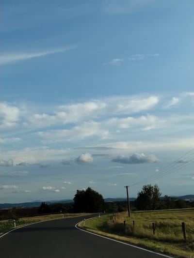 The image shows a rural road curving through a landscape with rolling hills and fields. The sky is a mix of blue with scattered white clouds. On the left side of the road, there is a field with dark soil, possibly a cultivated field, and some green vegetation. On the right side, there is a grassy field with a wooden fence and a telephone pole with power lines running across the frame. In the distance, there are trees and the silhouettes of hills. The road itself is asphalt with white markings. The overall impression is of a scenic, unpopulated rural area, likely in Velké Nepodřice, Czechia, given the context. There are no people or animals visible in the image.
