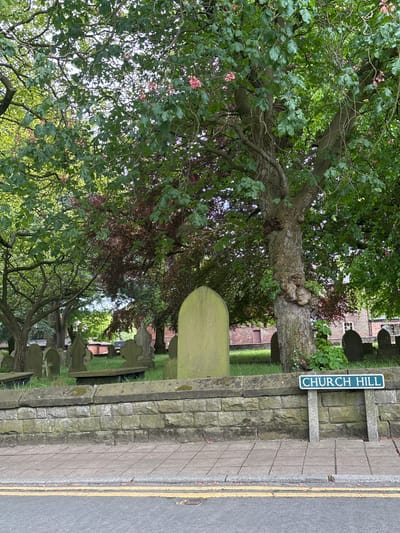 The image depicts Church Hill in Knutsford, United Kingdom, adjacent to a cemetery. A stone wall separates the cemetery from the sidewalk and road. A street sign reading "CHURCH HILL" is mounted on the wall. Trees, some with pink blossoms, overhang the cemetery and sidewalk. Tombstones of varying sizes and shapes are visible within the cemetery. The sidewalk is paved with gray blocks and the road has yellow lines marking the edge.