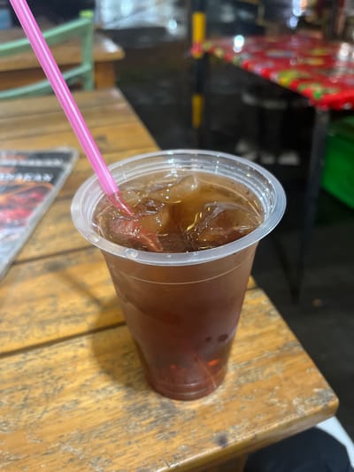 The image shows a close-up of a plastic cup filled with iced tea on a wooden table. A pink straw protrudes from the cup. In the background, a red and white checkered tablecloth is visible on another table, along with hints of green chairs. The scene appears to be outdoors, possibly at a street food stall or an outdoor cafe in Boja, Indonesia, given the context. The lighting suggests it may be evening or a dimly lit area. No people are clearly visible in the image.