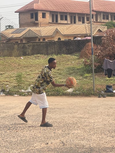A young male is depicted mid-stride on a coarse, light-colored asphalt surface in Umunekwu, Nigeria. He wears a patterned short-sleeved collared shirt, white shorts with blue trim, and dark grey slip-on shoes. His right hand holds a worn orange basketball extended forward and slightly downwards, with his left foot positioned ahead, suggesting an impending dribble or pass. Behind the individual, a chain-link fence separates the foreground from a grassy area containing sparse vegetation and scattered white plastic litter. In the background, beyond the fence, a lower single-story building with multiple yellow-tiled gabled roofs and rounded arch windows is visible, with solar panels installed on one roof section. Above and behind this, an unfinished two-story concrete block structure with a red-tiled roof and open window frames stands. To the far right, a pole is present, and a grey shirt and dark pants hang from an unseen support, with a dark backpack and sandals resting on the ground nearby. A reddish-brown dirt mound is situated behind the pole.