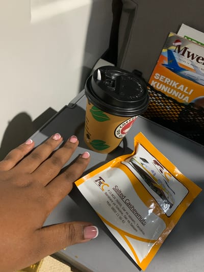 The image shows a close-up of a person's hand with manicured fingernails resting on a gray tray. Next to the hand is a disposable coffee cup with a black lid and a brown wrapper featuring green leaf designs. To the right of the coffee cup, a magazine is partially visible, with the word "Mweru" and "SERIKALI KUNUNUA" on its cover. Below the magazine and coffee cup is a sealed packet of "Salted Cashewnuts" with contact information for Tanzania Railways Corporation. The overall scene appears to be on a tray, possibly in a mode of transportation, given the presence of a magazine and snacks. The lighting suggests an indoor environment with overhead light casting shadows. The city of Magindu, Tanzania, is not explicitly visible or suggested by any elements in the image.