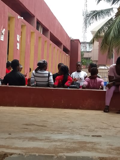 An outdoor scene in Umuokpu, Nigeria, depicts a group of approximately nine individuals gathered in front of a building. The dominant structure on the left is a two-story building featuring reddish-brown upper walls and lower sections with prominent vertical yellow columns or panels. Several white papers are affixed to the reddish-brown wall. An air conditioning unit is visible in an upper window recess, along with white light fixtures mounted on the wall.
In the midground, a low, reddish-brown concrete barrier or seating area extends horizontally across the frame. Behind this barrier, a group of people, primarily young adults, are positioned. From left to right: one person in a red top is partially obscured; one individual in a black shirt faces away from the viewer; one person wears a grey and white striped shirt and a patterned head covering, facing away; one person in a dark shirt is partially obscured; one person in a red t-shirt with black designs faces away, with a black bag and a blue bottle nearby; a central figure in a white "CK" branded t-shirt faces right, appearing to speak; two individuals in light pink tops are partially visible behind the speaker; and on the far right, one person in a light pink long-sleeved top and matching leggings is seated on the barrier, with a blue plastic water bottle next to them and black sandals on their feet.
The foreground consists of a light brown, uneven concrete or dirt surface. In the background, to the right of the building, a tall metal lattice communication tower is visible against a bright, overcast sky. Portions of another building with a light-colored roof and walls, and green palm fronds from a tree, are also present. The scene is illuminated by natural daylight.