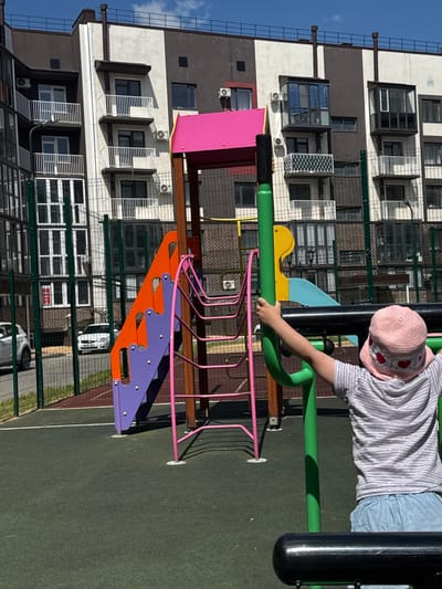 The image shows a child in a pink hat and a striped shirt playing on a colorful playground. The playground has bright pink and purple plastic steps, orange accents, and green metal bars. The child's hand is gripping a green metal bar. In the background, there is a modern apartment building with balconies and air conditioning units. The sky is blue and appears clear, suggesting a sunny day. The playground is situated on a green, soft surface, likely for safety. The location is identified as Вязники, Russia. The image is taken from a slightly elevated, angled perspective, capturing both the child and the surrounding environment.