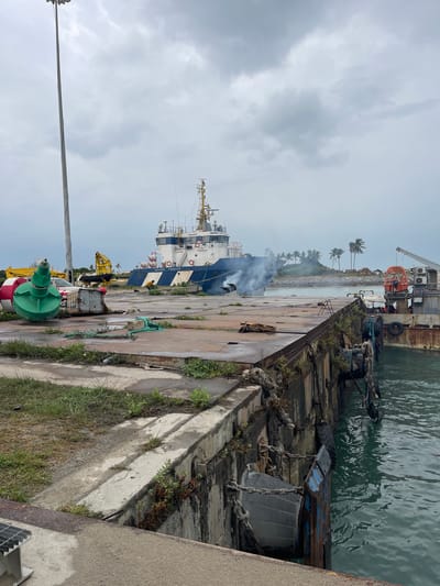 The image depicts a waterfront scene, likely a port or dock area, possibly in Ebute Leki, Nigeria. A large, blue and white ship is docked alongside a concrete quay. Smoke is visible near the ship, possibly emanating from a smokestack. The quay is lined with various pieces of equipment, including a crane, possibly a few buoys, and other maritime-related objects. The quay itself shows signs of wear and age, with concrete and metal elements present. Palm trees are visible in the background. The sky is overcast.