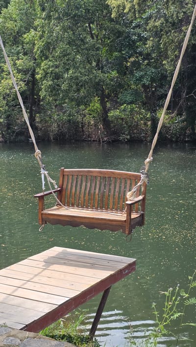 The image shows a wooden swing hanging over a body of water near a wooden dock. Lush green trees line the far bank. The location is Nam Ninh Hoa, Vietnam.