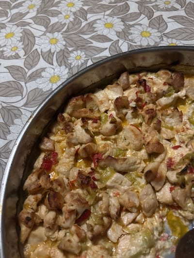 The image shows a close-up, overhead shot of a metal baking dish containing a chicken dish. The chicken appears to be cut into bite-sized pieces and mixed with a creamy sauce, including green and red peppers. The dish is on a tablecloth, featuring a floral pattern of white daisies with yellow centers and gray leaves on a light background. No people are visible in the image. The location of Rakitovo, Bulgaria is extraneous and does not appear to be relevant to the image's contents.